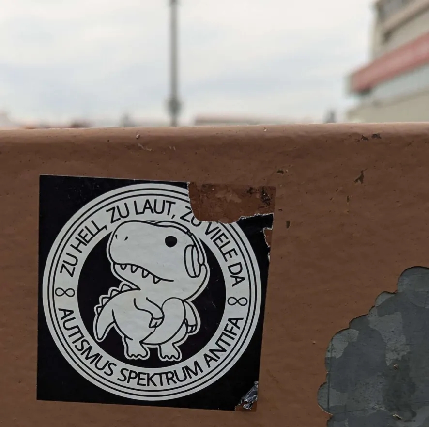a sticker somewhere in public. it has a dino with headphones on it and the text 'zu hell, zu laut, zu viele da - autismus spektrum antifa' which translates roughly to 'too bright, too loud, too many people - autism spectrum antifa'